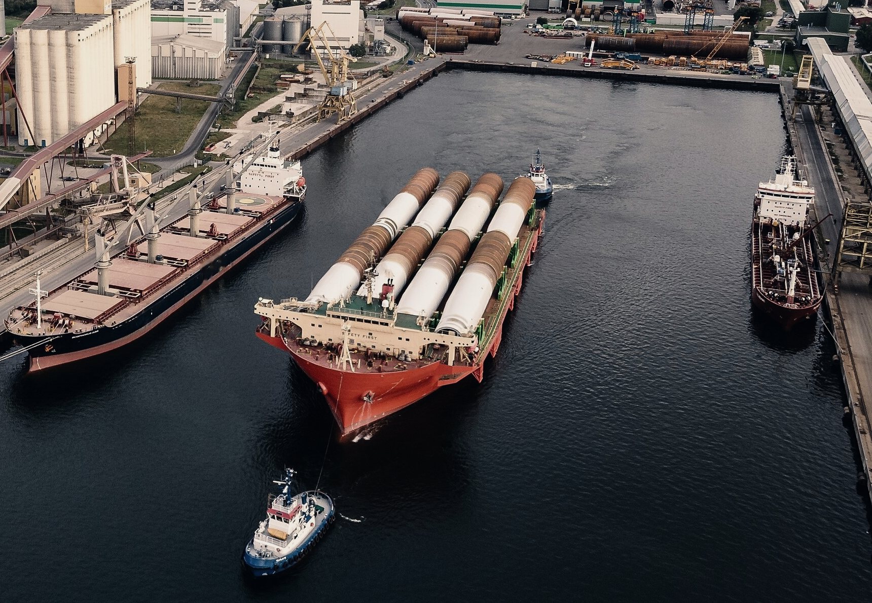 Aerial view of a heavylift deck carrier loaded with eight wind turbine monopiles, leaving a port basin escorted by tugs