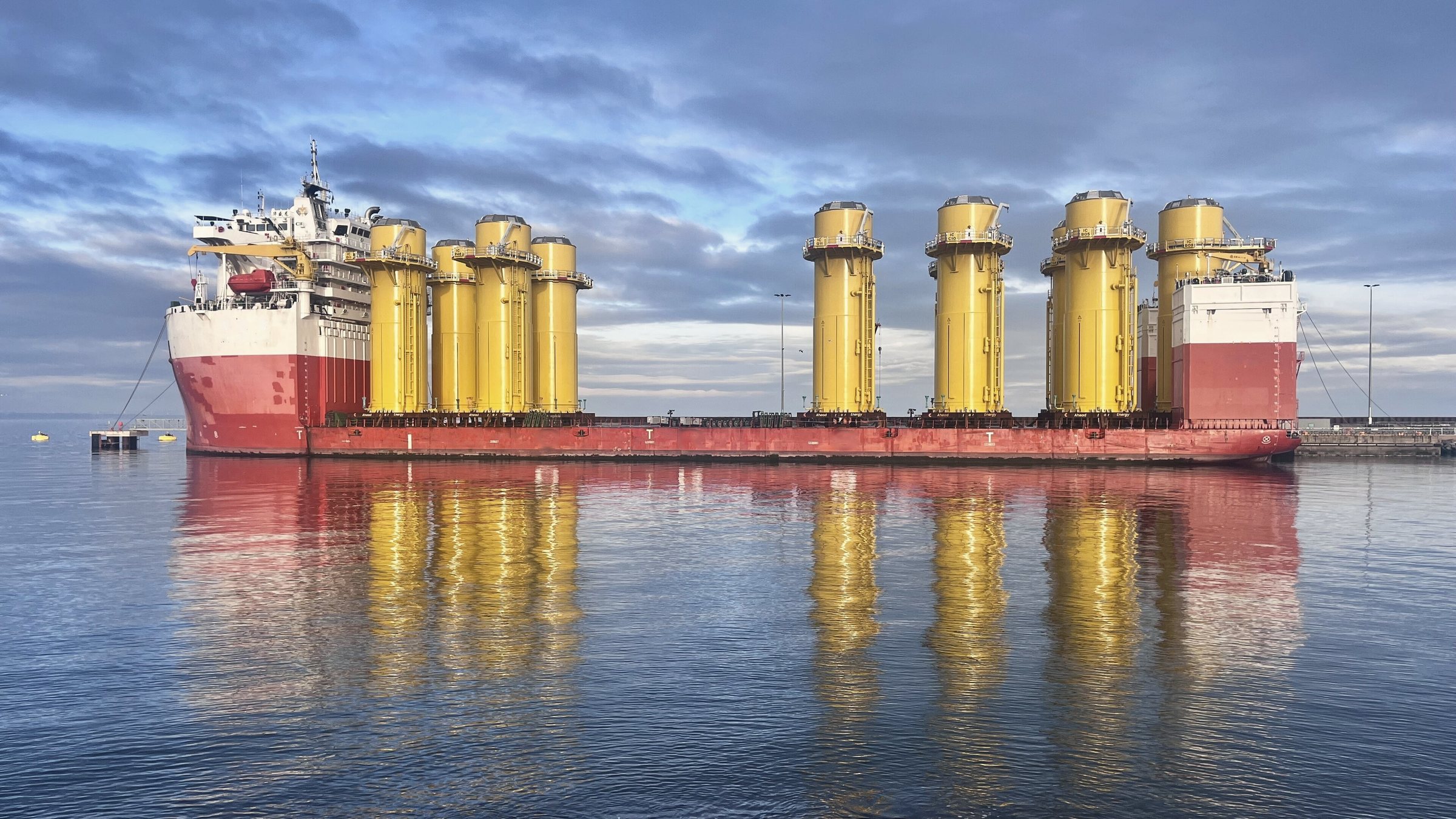A semi-submersible heavy transport vessel loaded with yellow transition pieces for an offshore wind farm, mirrored in still water
