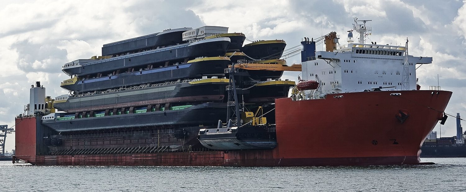 A semi-submersible vessel carrying a stack of river barges, pontoons and workboats on its deck