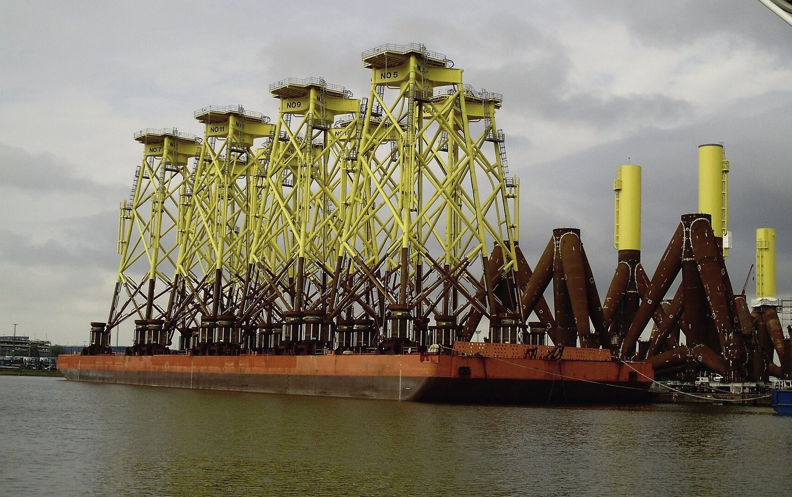 A row of yellow lattice jacket foundations standing on an orange deck barge